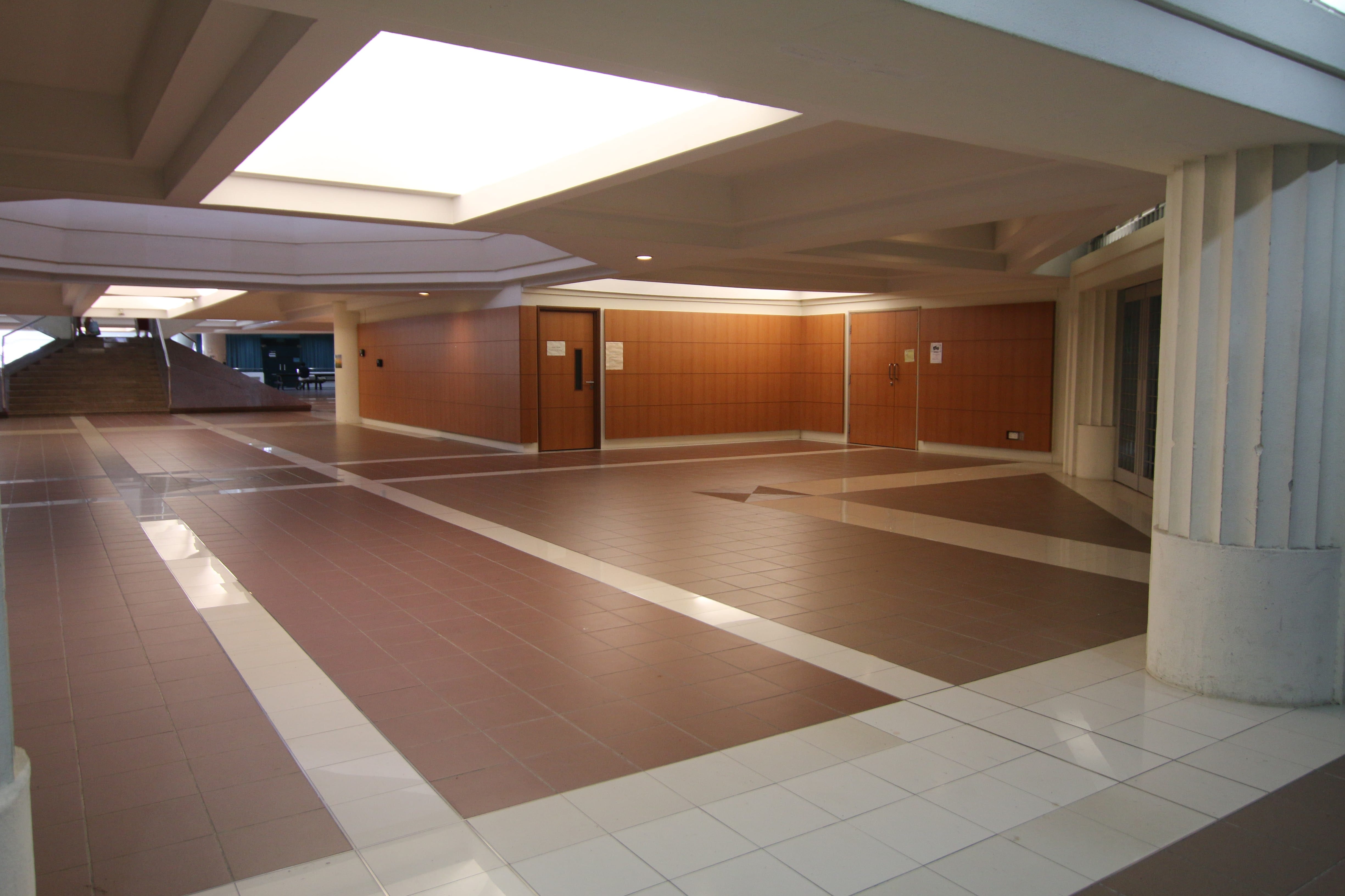Empty indoor lobby with brown and white tiled floor, white columns, warm-lit walls, and a staircase in the background.